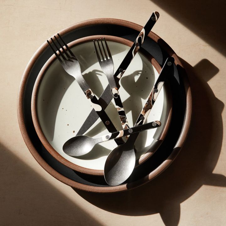 A group of black, white and cream cutlery (salad fork, dinner fork, knife, soup spoon and teaspoon) stacked on white and black bowls