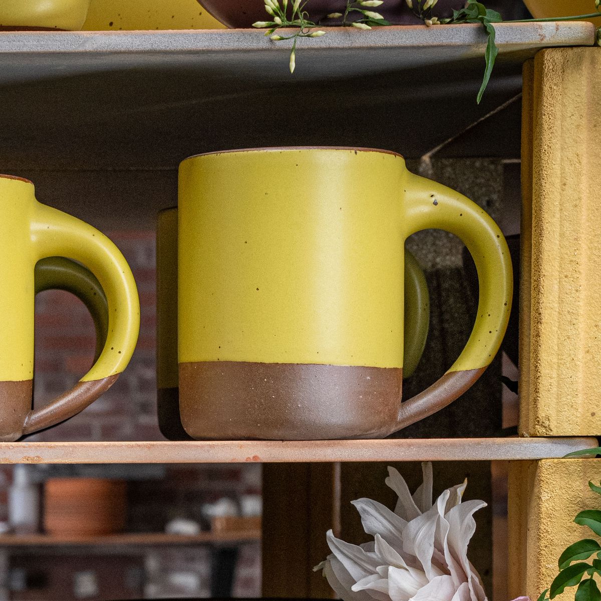 A large sized ceramic mug with handle in a zesty yellow color featuring iron speckles and unglazed rim, sits on a wood shelf