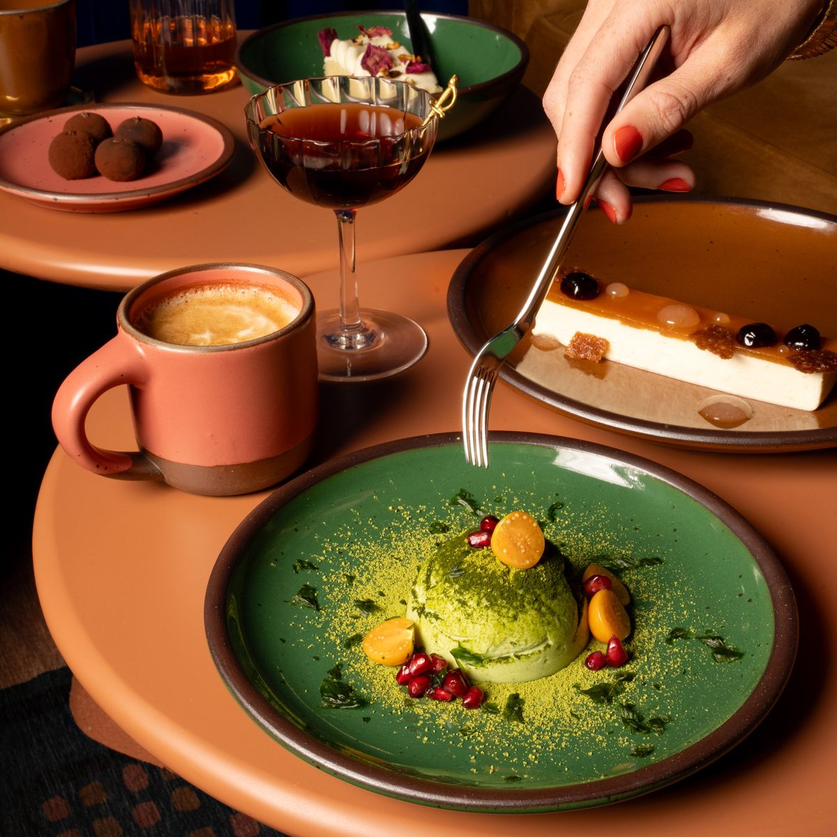 A hand holds a fork over an artful dessert plated on a medium sized ceramic plate in a glossy emerald green color, surrounded by other ceramic plates and mugs in watermelon pink, glossy brown, and glossy green colors.