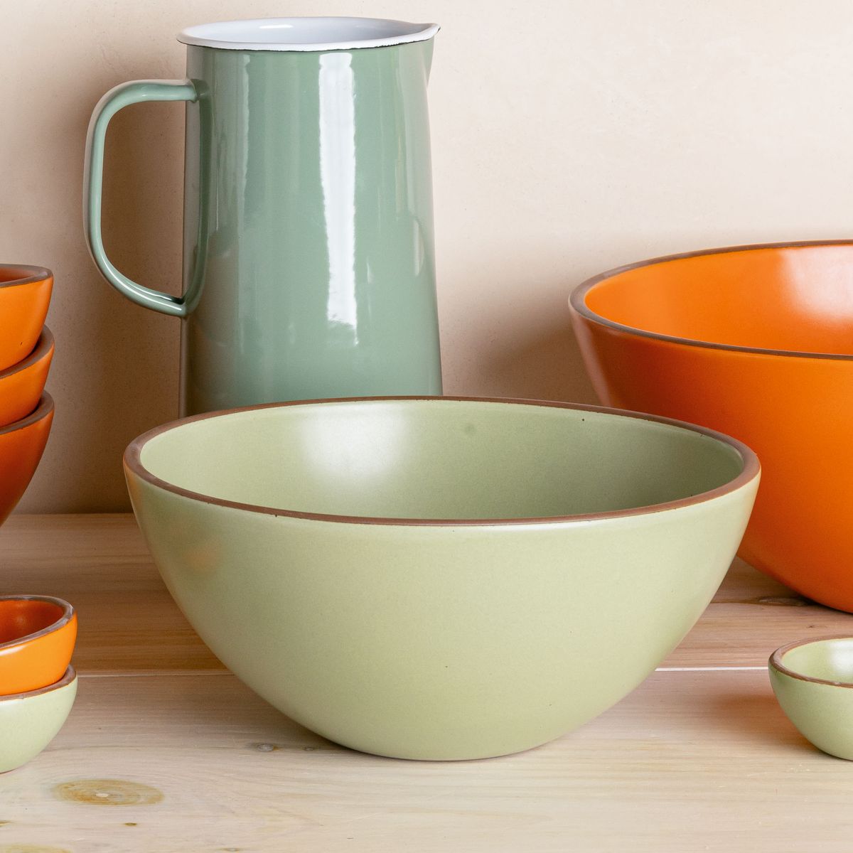 On a shelf, sits a calming sage green popcorn bowl with a sage green watering pitcher and other green and bold orange pottery.