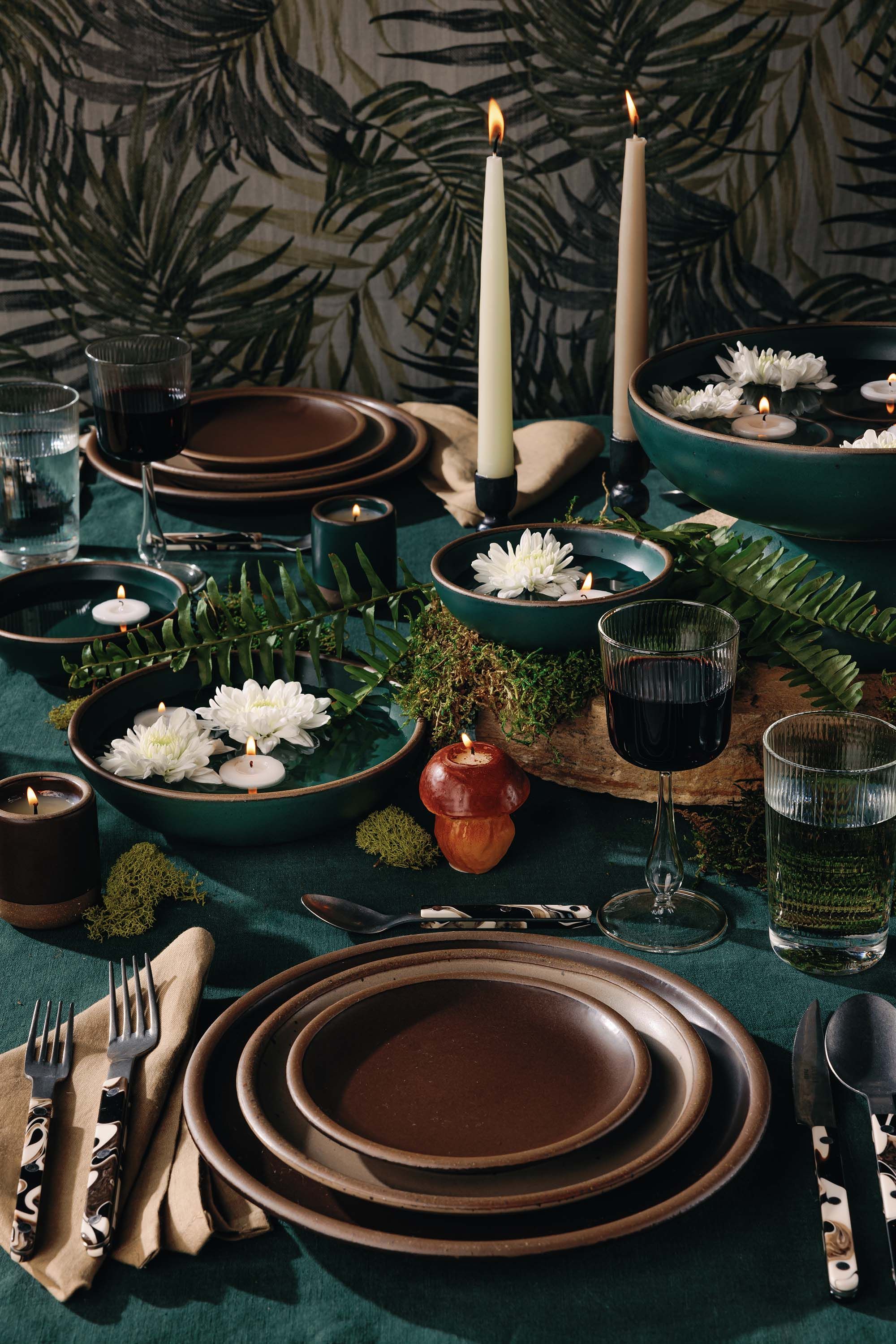 A tablescape scene featuring brown and teal ceramic plates and bowls, lit tapered candles, ferns, white flowers, with sophisticated flatware and table napkins. All on a dark teal tablecloth with a fern wallpaper background
