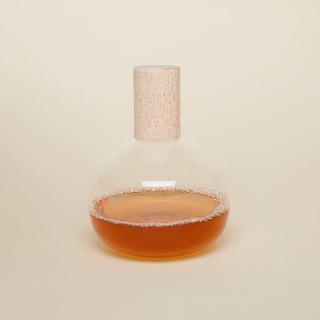 A hand holds a short glass bottle with wide body and a cylindrical wooden top, filled with vinegar