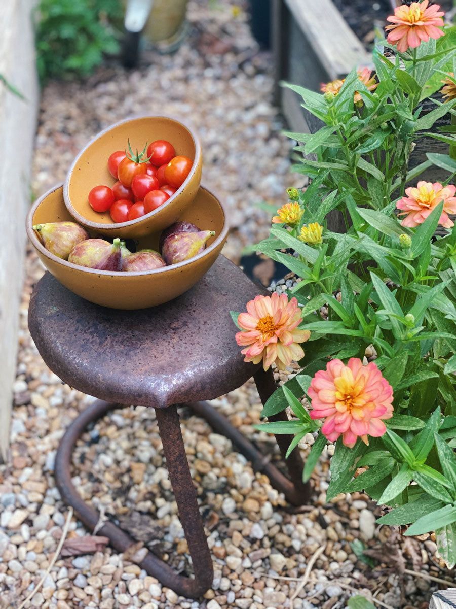 Outside, two ceramic bowls are filled with freshly picked vegetables and fruit, sitting on an iron stool next to a flower bush