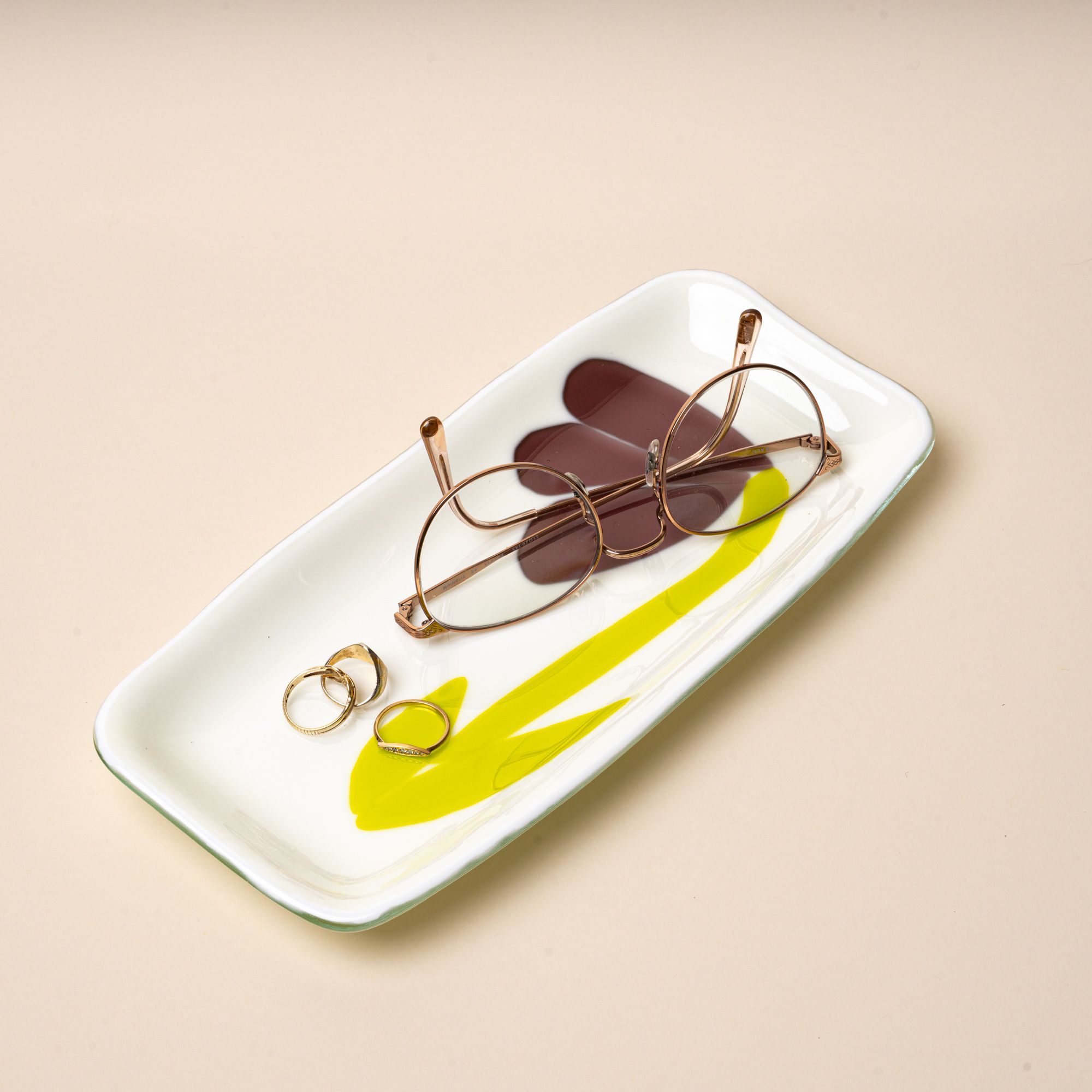 Glasses and rings sit on a white rectangular glass catchall tray with a design featuring a minimal purple flower with green stem.