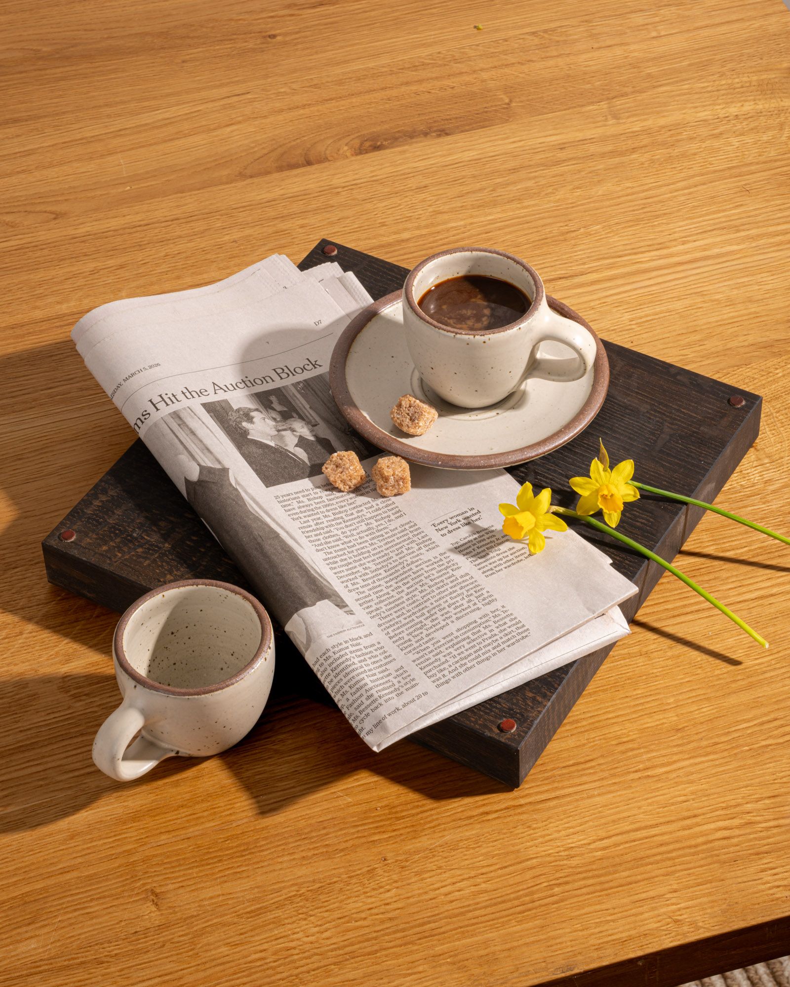 Small stoneware espresso cup with handle and saucer, in a warm off-white color with iron speckles, styled with another espresso cup, newspaper, and flower stems.
