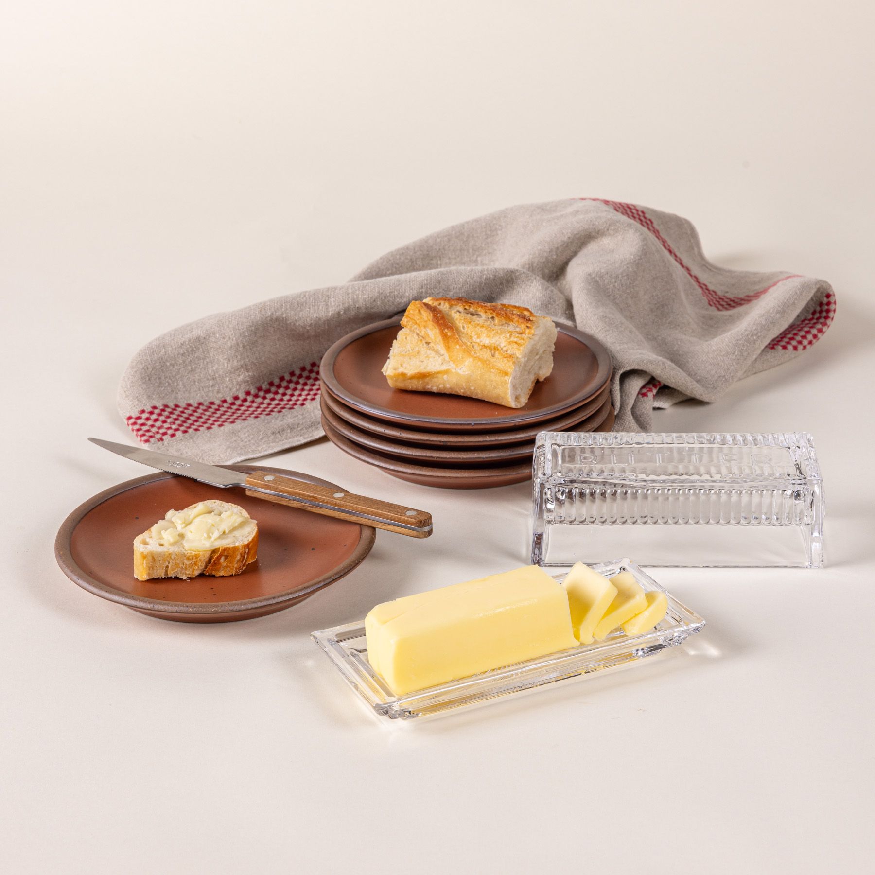 Traditional glass butter dish with 'Butter' on top and butter inside. Off to the side are terracotta plates with bread and a knife.