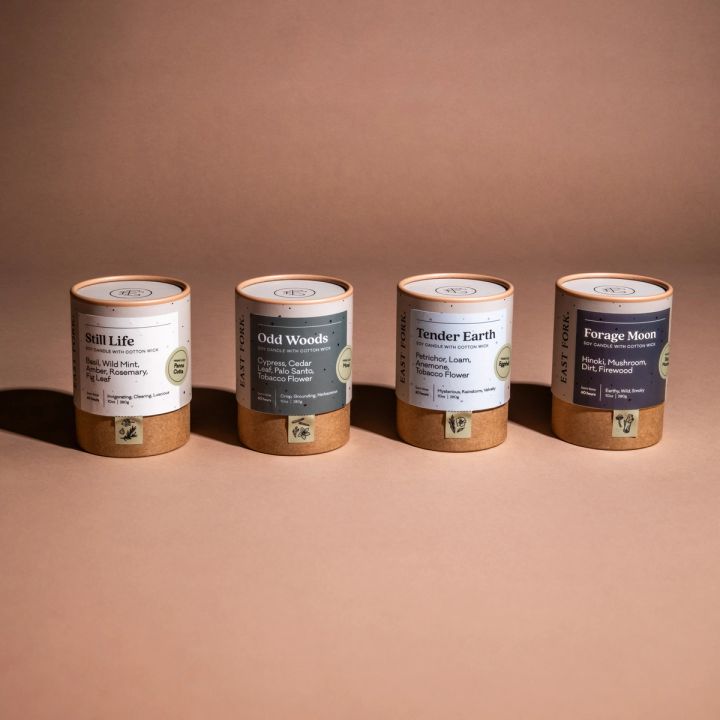 4 small candles in their cardboard packaging sitting together in a line. All the labels are in neutral colors and read "Still Life", "Odd Woods", "Tender Earth", and "Forage Moon"