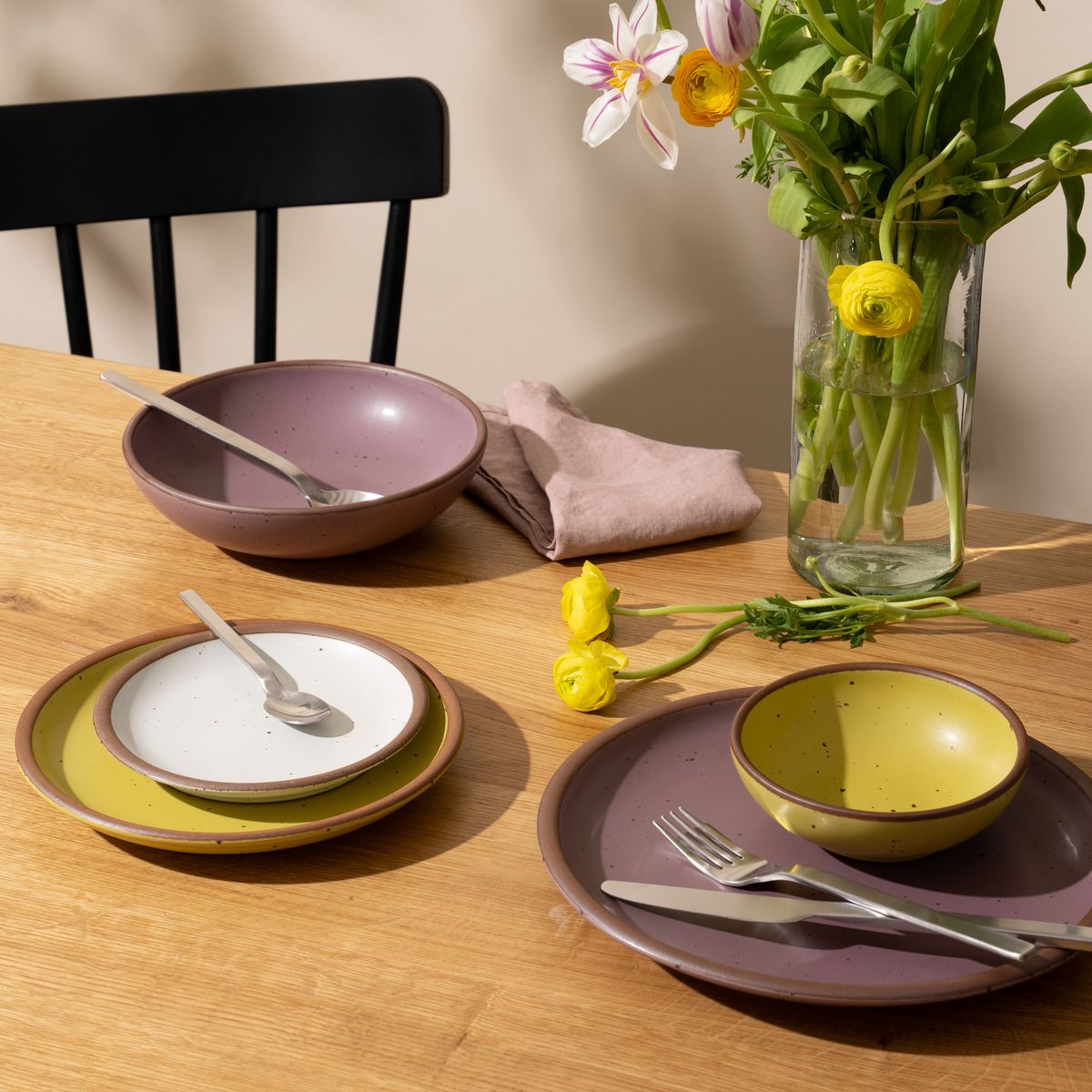 An artful dinnerware set featuring 3 different sized plates and 2 small and large shallow bowls in soft mauve purple, zesty yellow and cool white colors on a table. There is also silverware on the plates, a tall glass of water, and a tall clear vase with yellow flowers.