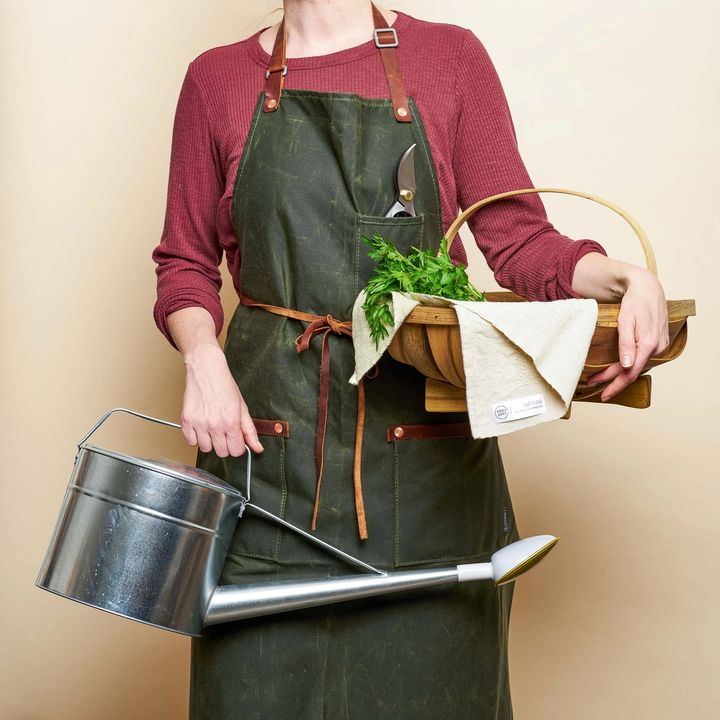 A gardener ready to get dirt under their nails - wearing a waxed apron, holding a water can, and a wicker basket.