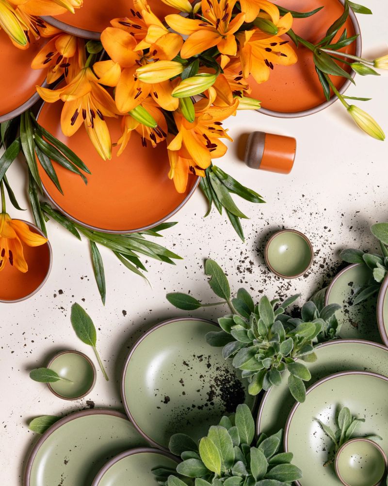 An artful overhead view of ceramic plates, bowls, and cups in a bold orange and calming sage green surrounded by daylilies and lamb's ear. The bold orange pottery and daylilies are on the left, the sage green pottery and lamb's ear on the right.
