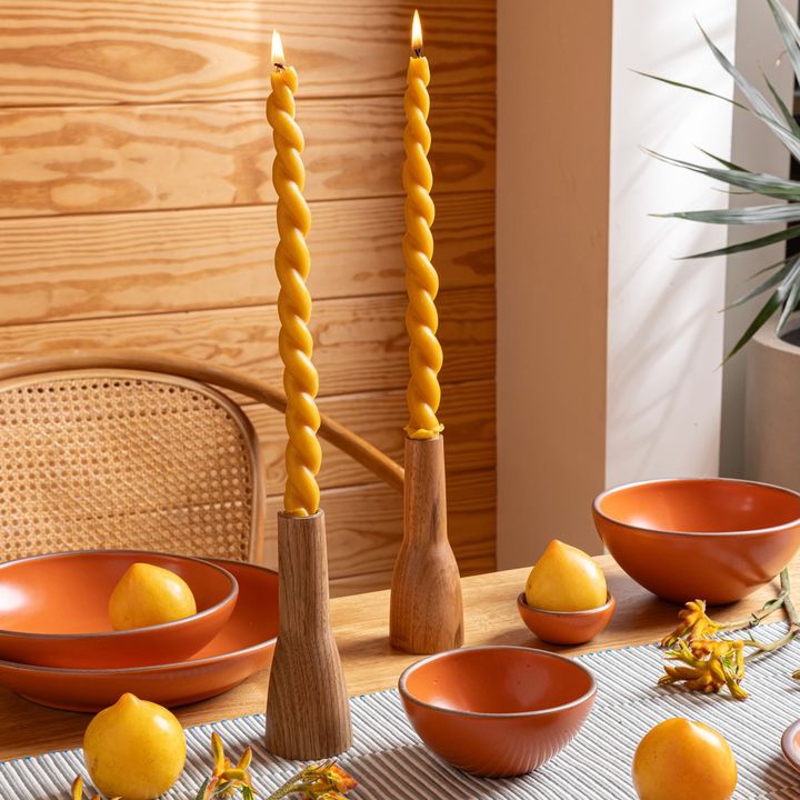On a table are two twisted beeswax candles in two wooden candleholders. Surrounding the tapers are orange bowls, lemons, and greenery.