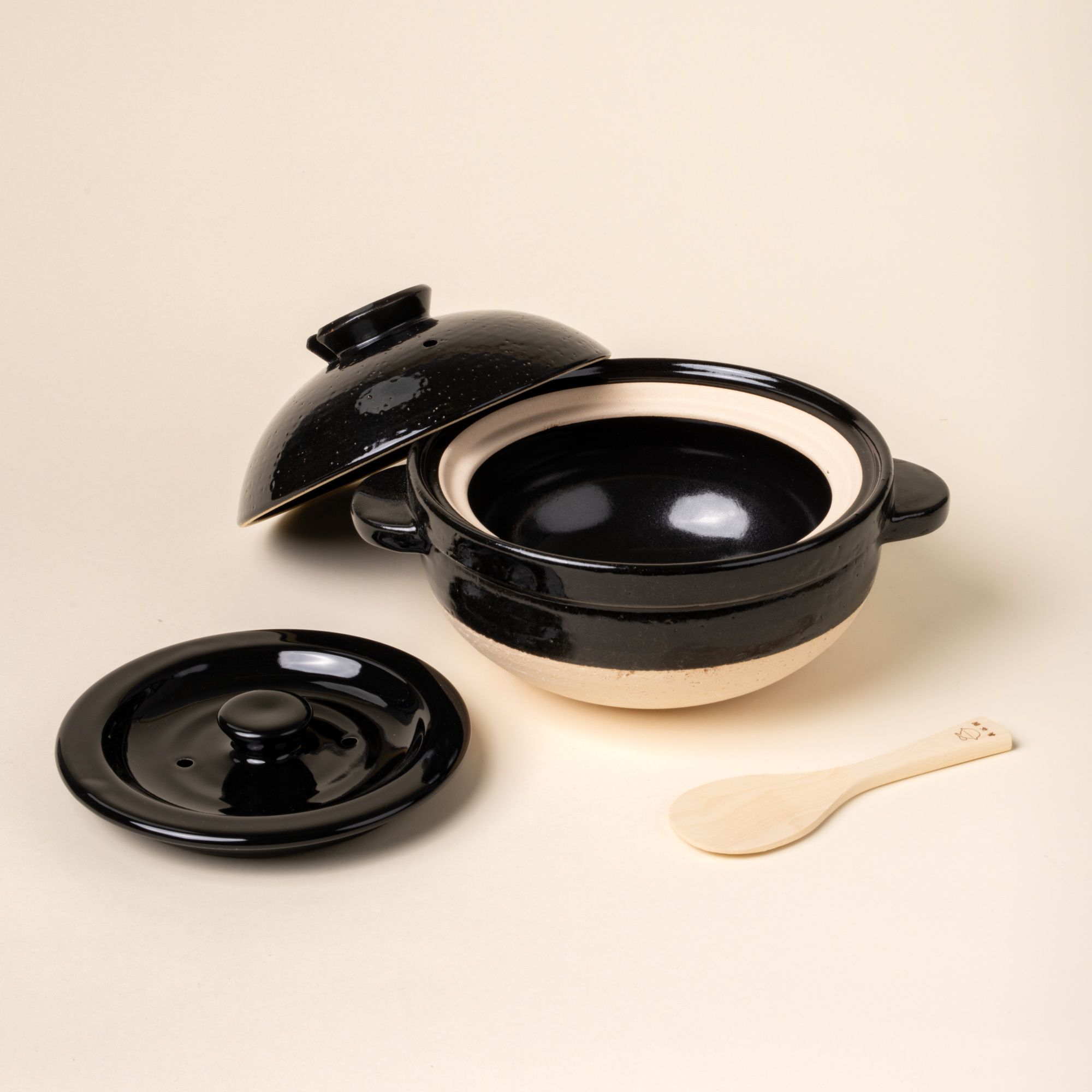Black ceramic donabe pot with two lids and a small wooden spoon on a light background.