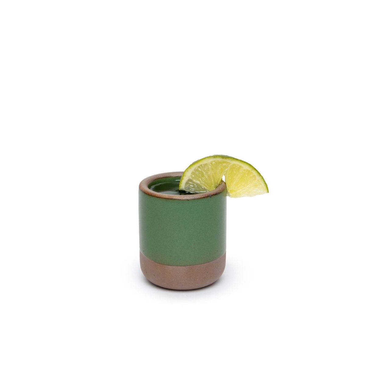 A small, short ceramic mug cup in a deep, verdant green color featuring iron speckles and unglazed rim and bottom base, filled with a drink and topped with a lime slice