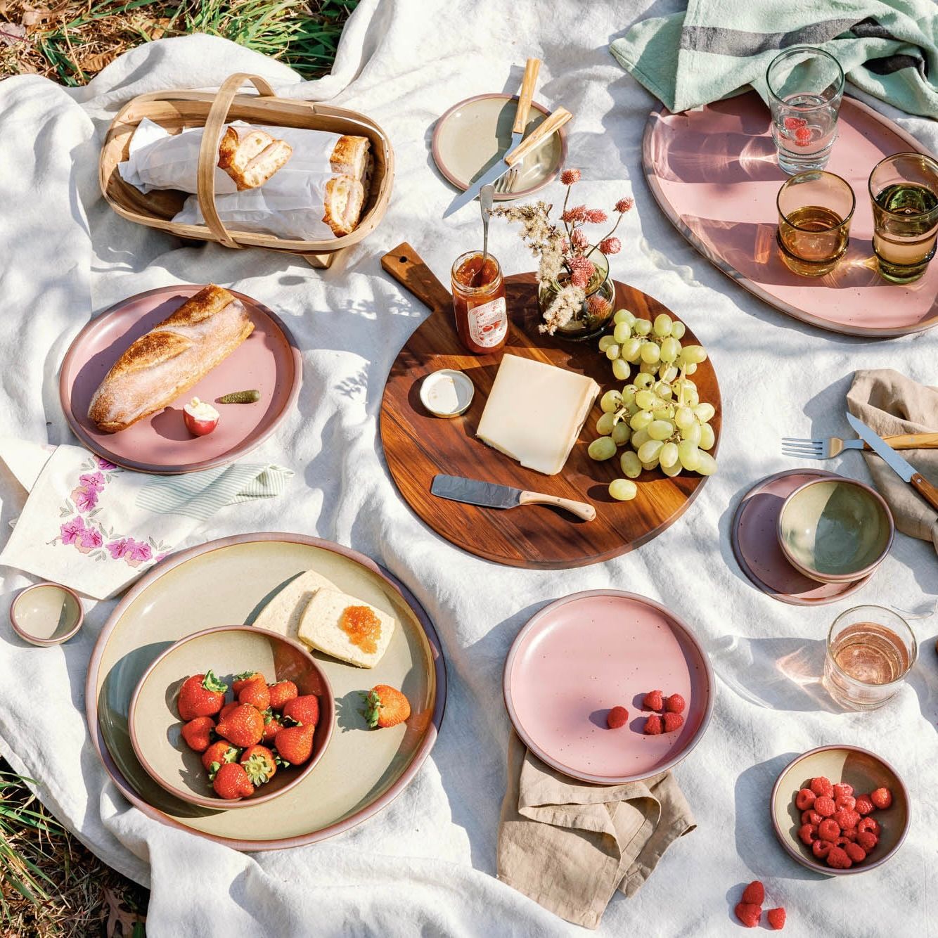An outdoor picnic filled with ceramic dinnerware in dusty mauve pink and grey-green colors, bread, berries and cheese, a charcuterie spread, glassware and more.