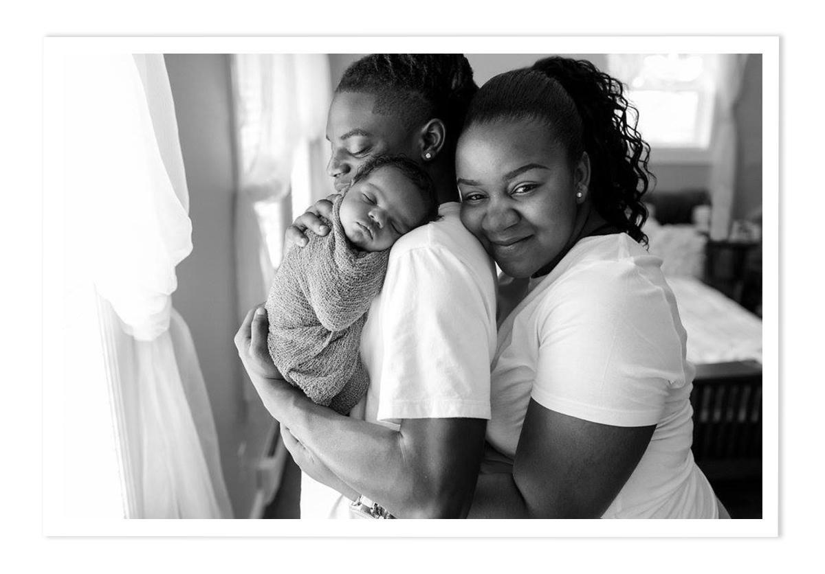 A black and white photo of 2 people holding each other facing the left and smiling, the person to the left is holding a newborn baby