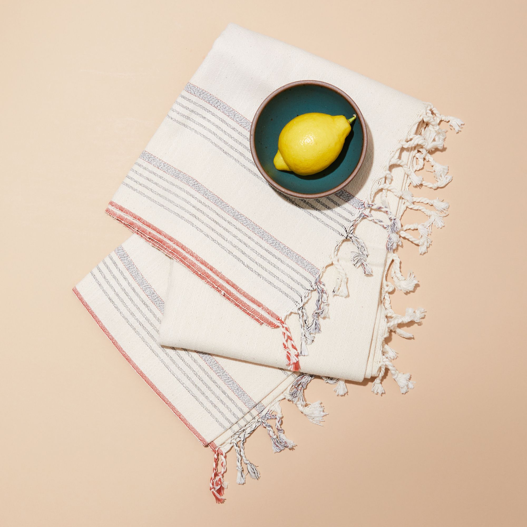 A bowl with a lemon resting on cream-colored cotton Turkish towel with blue and coral stripes and tassels 