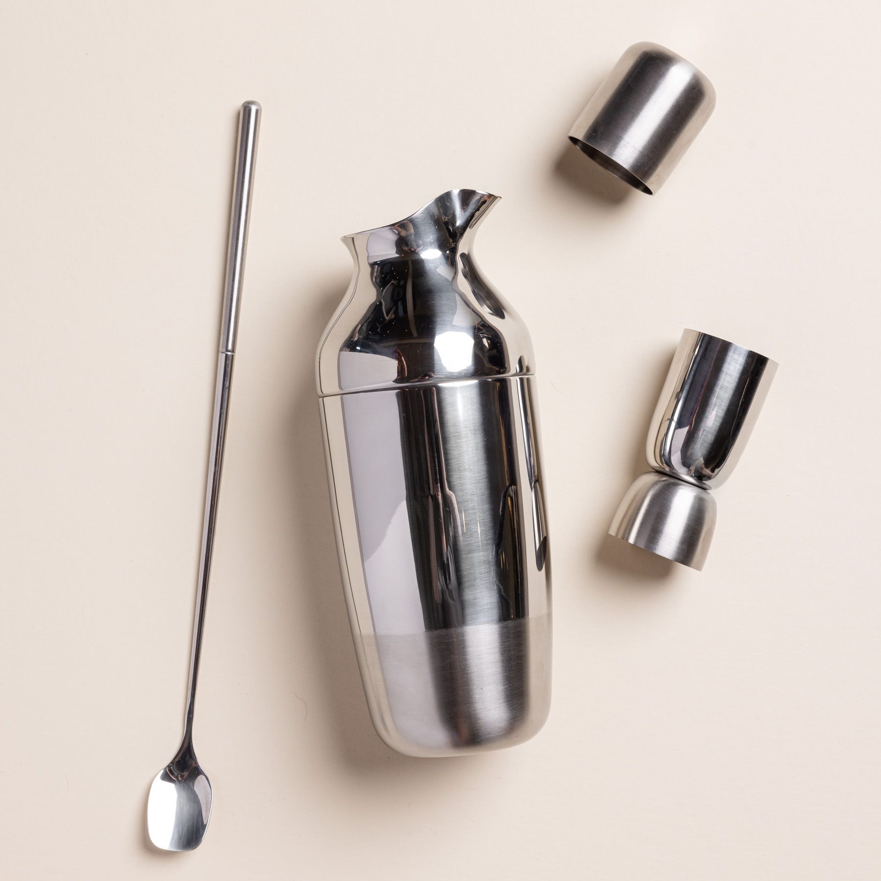 The Mescola Cocktail Set (including a stainless steel shaker, jigger, and cocktail spoon) laying flat on an off-white surface