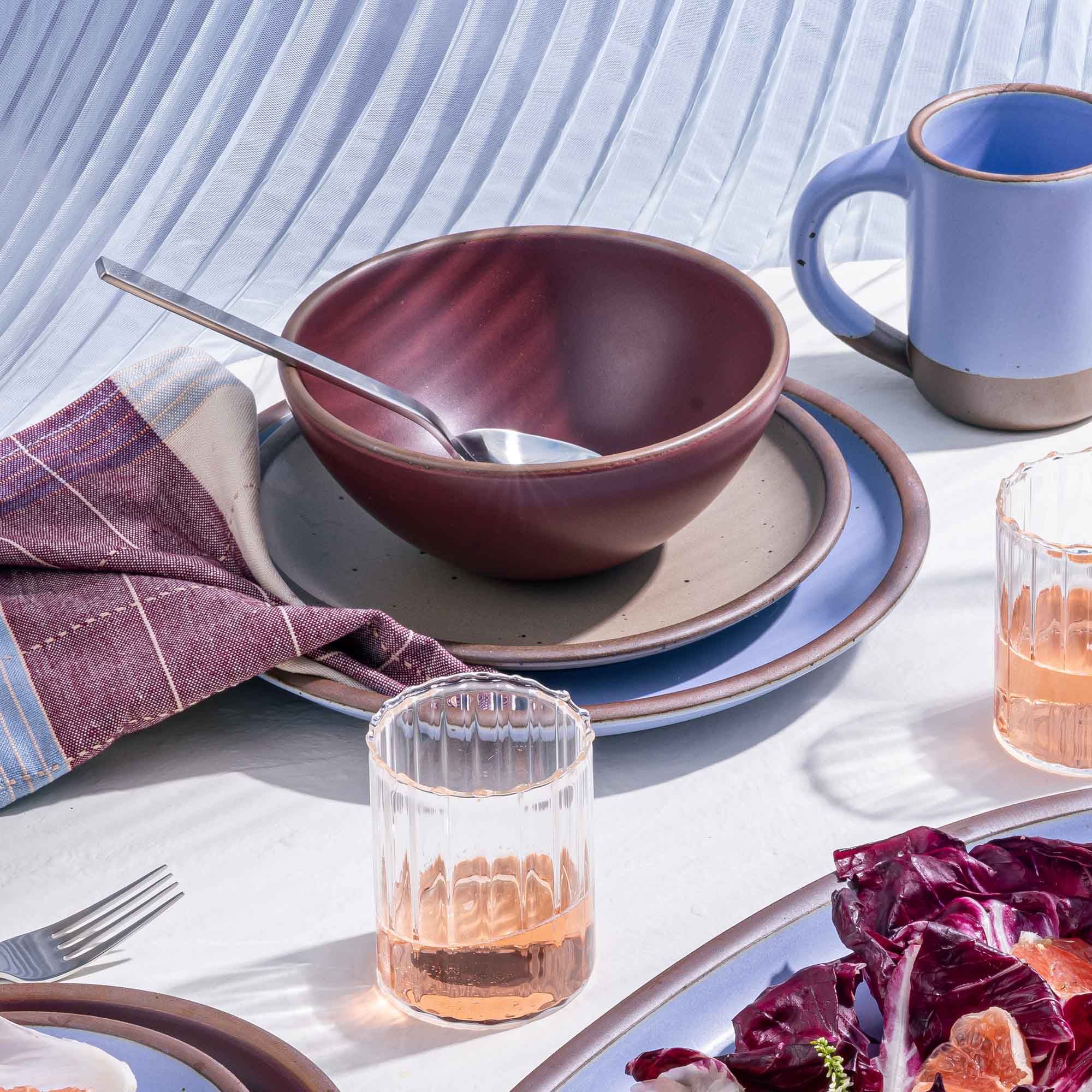 A rounded soup bowl in a plum color with a spoon, napkin, other dishes, and fluted glassware