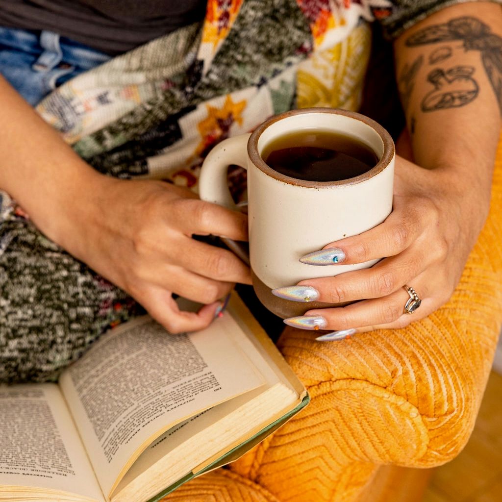 A person is sitting in a yellow chair reading holding an East Fork ceramic mug in an off-white color