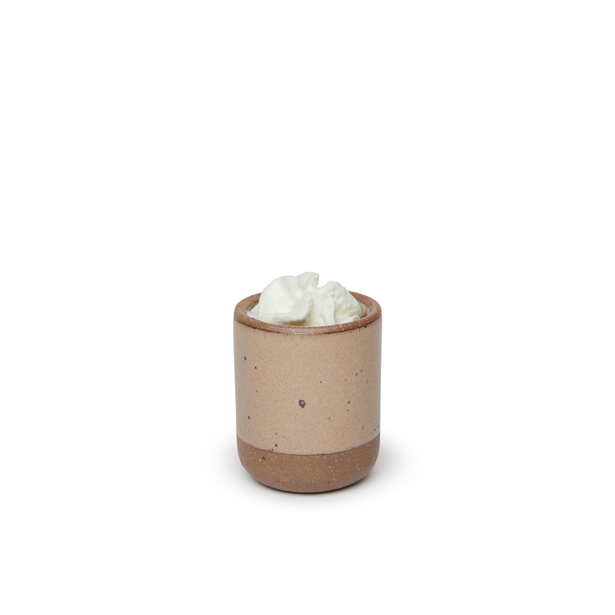 A small, short ceramic mug cup in a warm pale brown color featuring iron speckles and unglazed rim and bottom base, with whipped cream on top.
