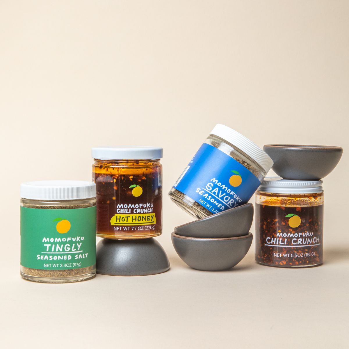 4 little ceramic bowls in a soft cool grey color surrounded by 4 jars of Momofuku seasoned salts and chili crunch.