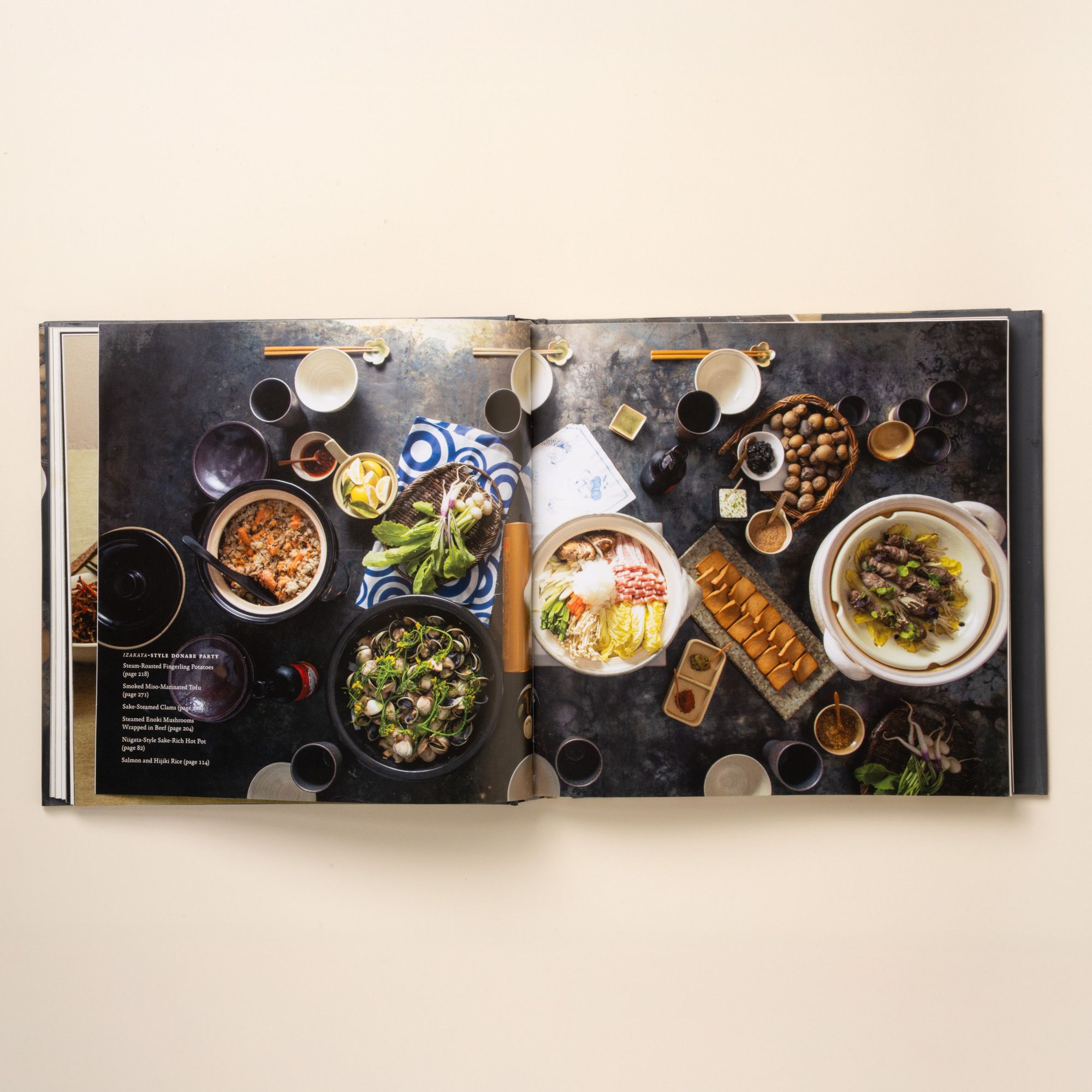 Open cookbook showing a Japanese-style dinner spread with bowls, plates, and shared dishes arranged on a dark tabletop.