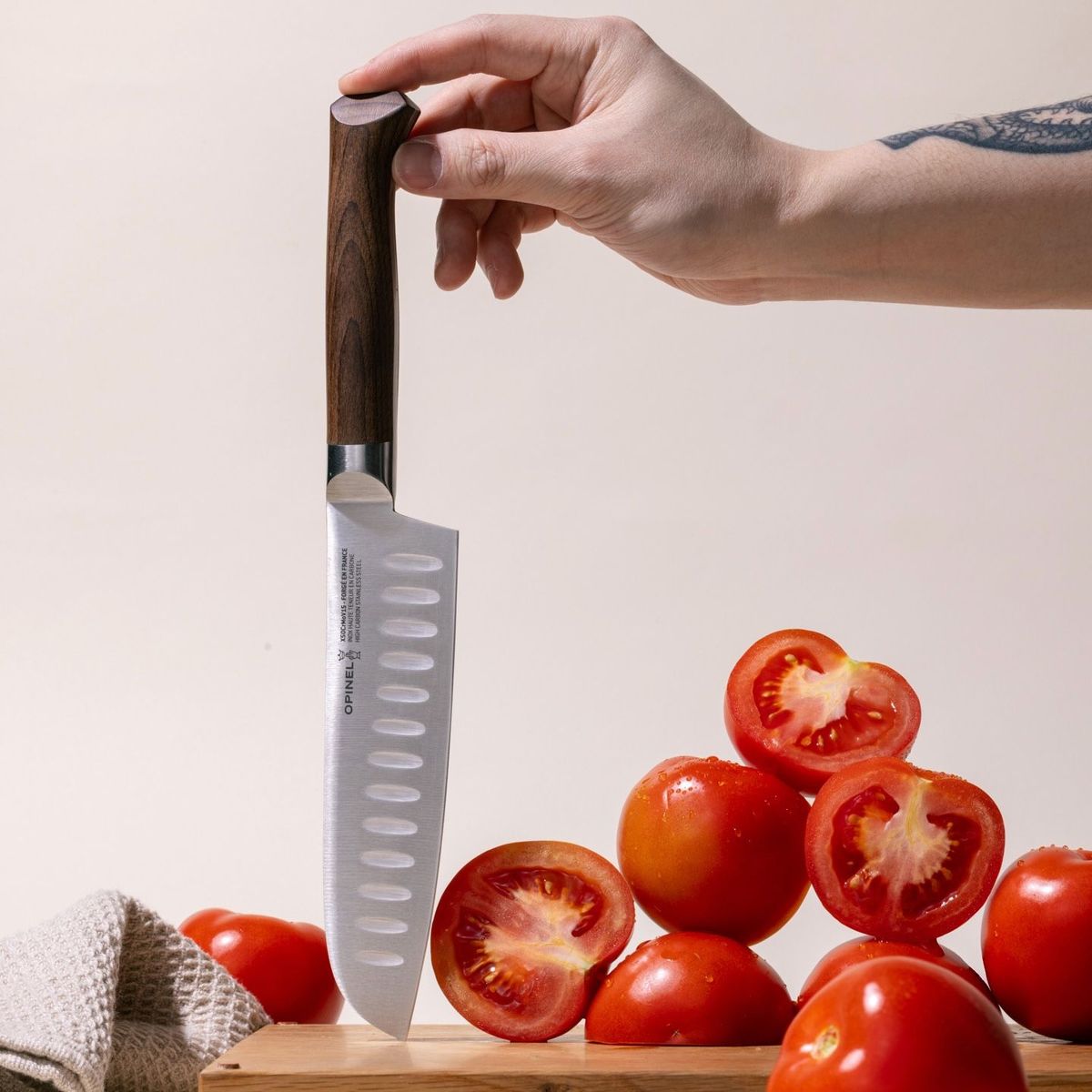 A hand holds up a santoku knife with a sharp steel blade with dimples and a beveled dark wood handle. The knife is being held upside down on a cutting board with a tower of tomatoes both full and sliced.