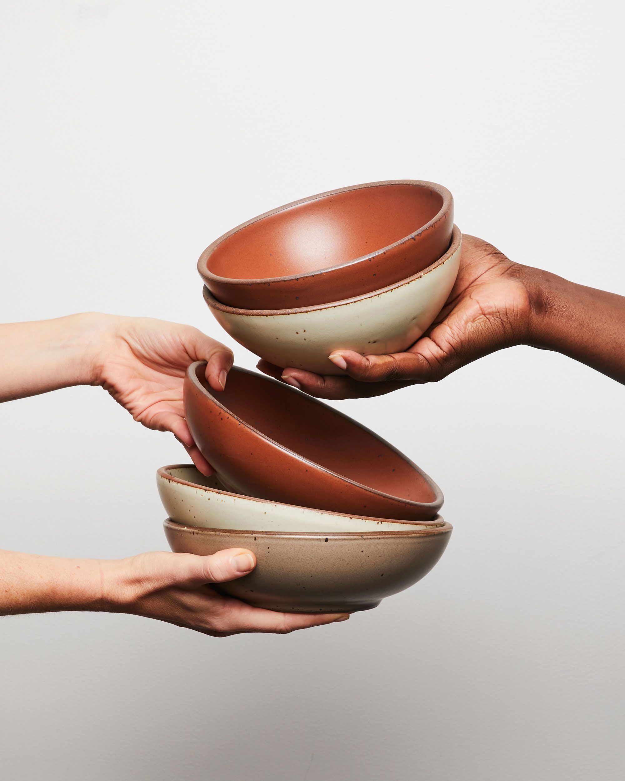 Hands hold out ceramic shallow bowls in terracotta, off-white, and warm pale brown colors.