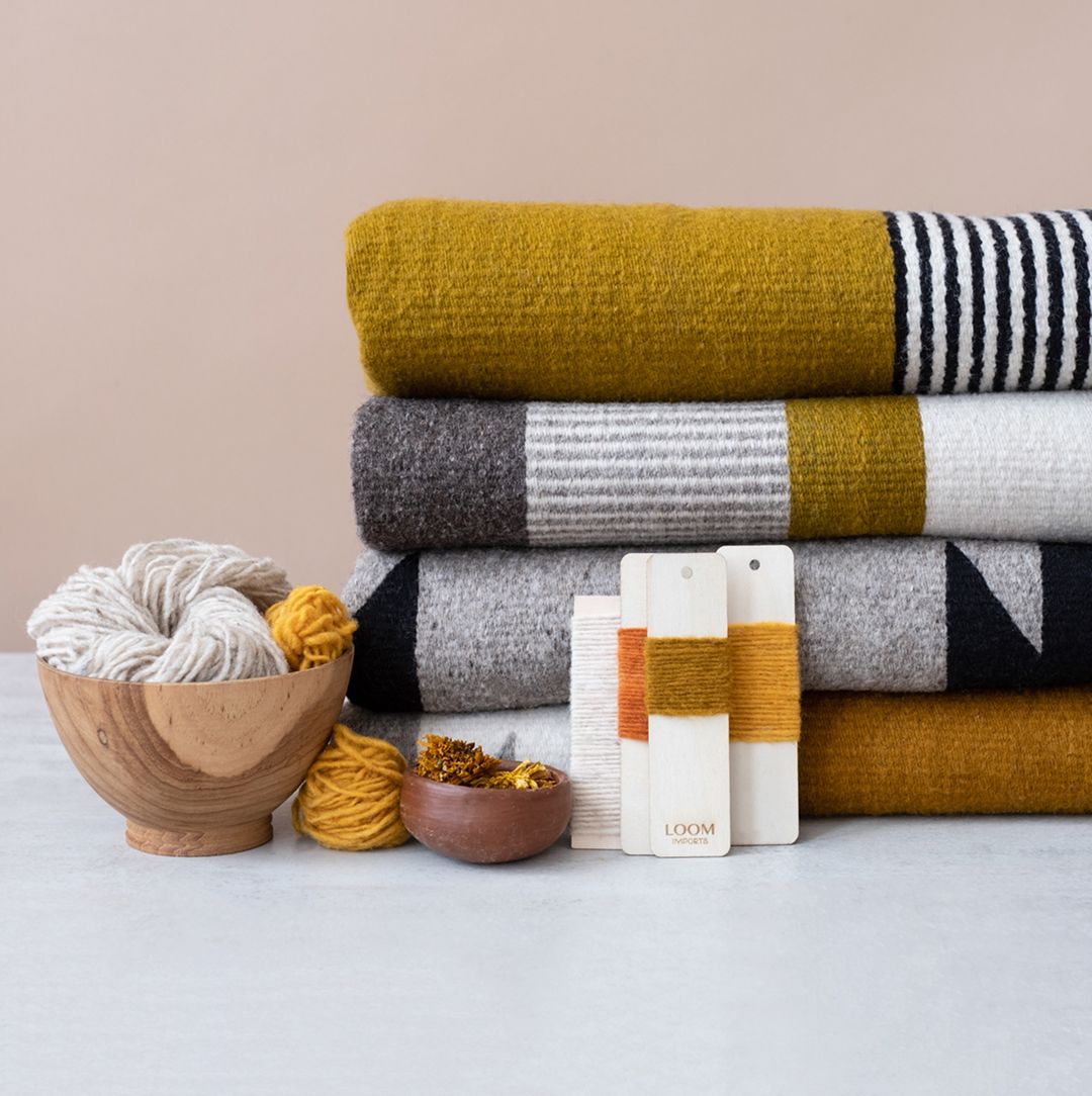 Stack of woven blankets in mustard yellow, gray, and black with yarn balls, wooden bowls, and Loom Imports workshop swatches.