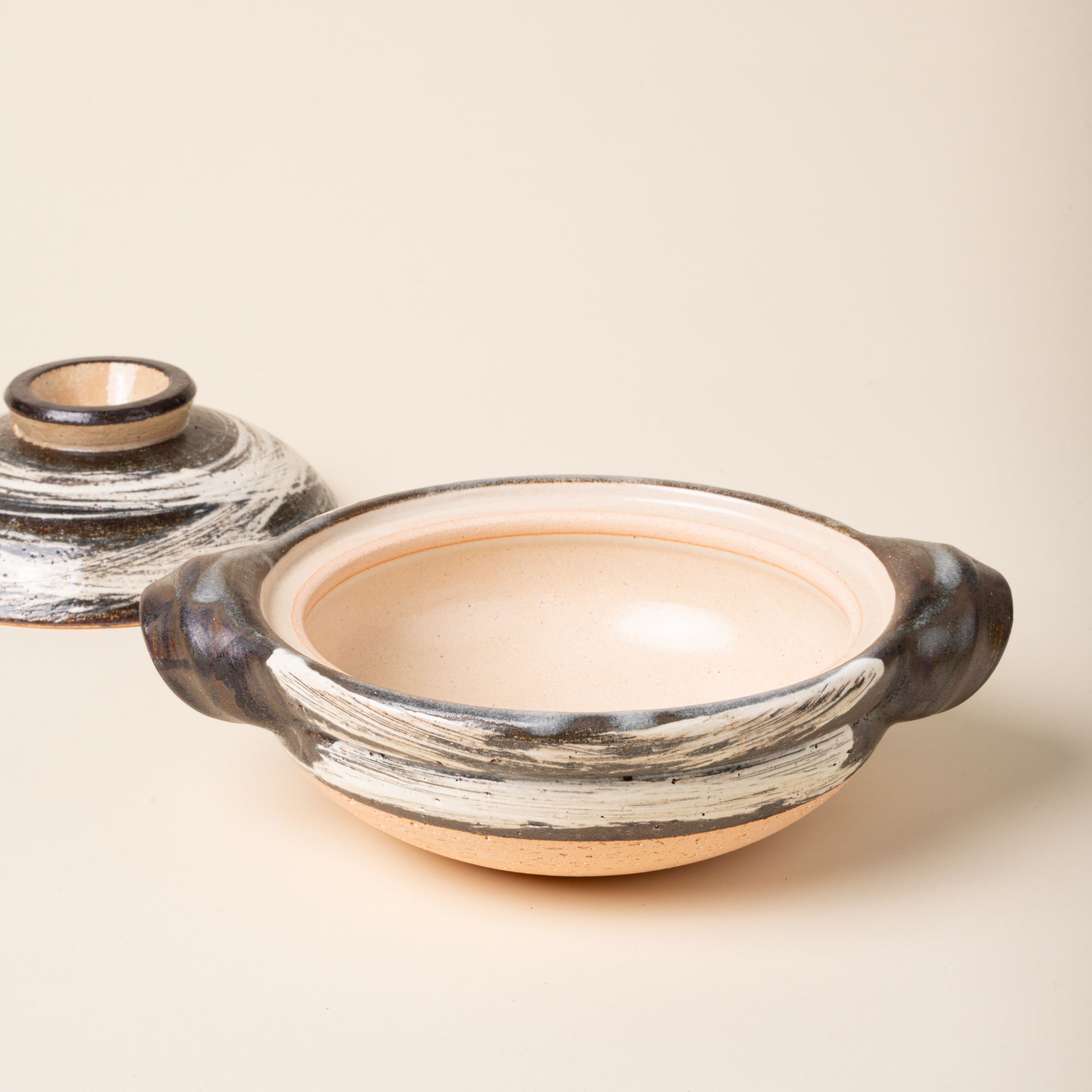Shallow ceramic donabe with sculpted side handles and a brushed black-and-white glaze over warm natural clay, with the lid off to the side.