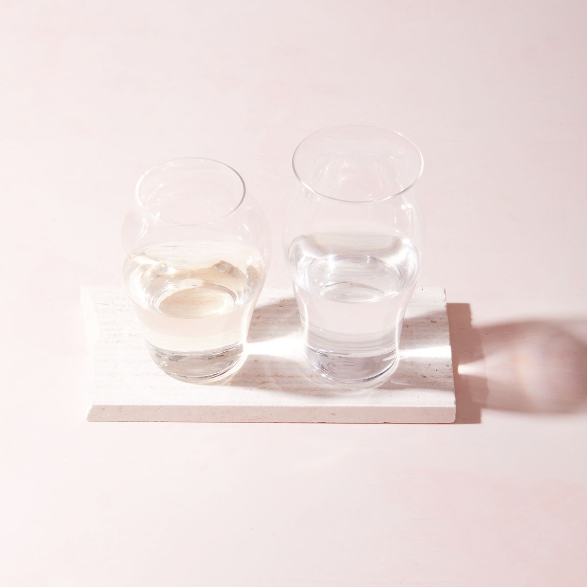 Two clear glasses filled with water, one taller than the other, both with curved bodies, sitting on a rectangular piece of wood