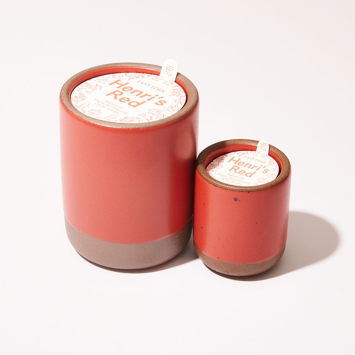 Small and large ceramic vessel next to each other in orange-red color with candles inside each. On top of each is a packaging label sitting that reads "Henri's Red"