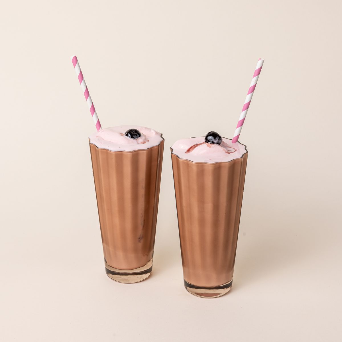 Two tall transparent light amber glasses with wide grooves on the side. The glasses are filled with a milkshake, cherry, and a striped pink and white straw