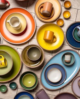 Ceramic dinnerware of plates, platters, bowls, mugs and cups are all artfully arranged close together all in different colors of the rainbow.