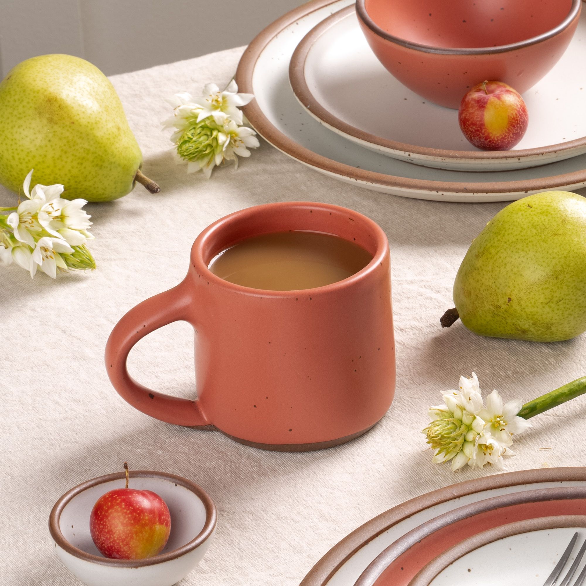 Coffee in a watermelon pink mug with pears, flowers, and stoneware plates on a neutral tablecloth.