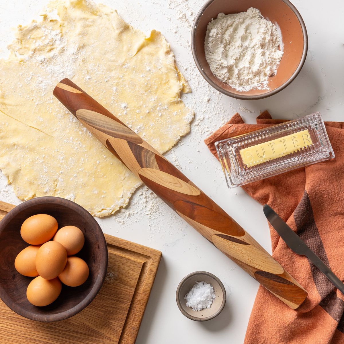 On a white countertop is a bunch of items in the middle of baking including a mixed wood rolling pin that tapers at the end, flat rolled out dough, a bowl of eggs, a bowl of flour, glass butter dish, and a napkin.