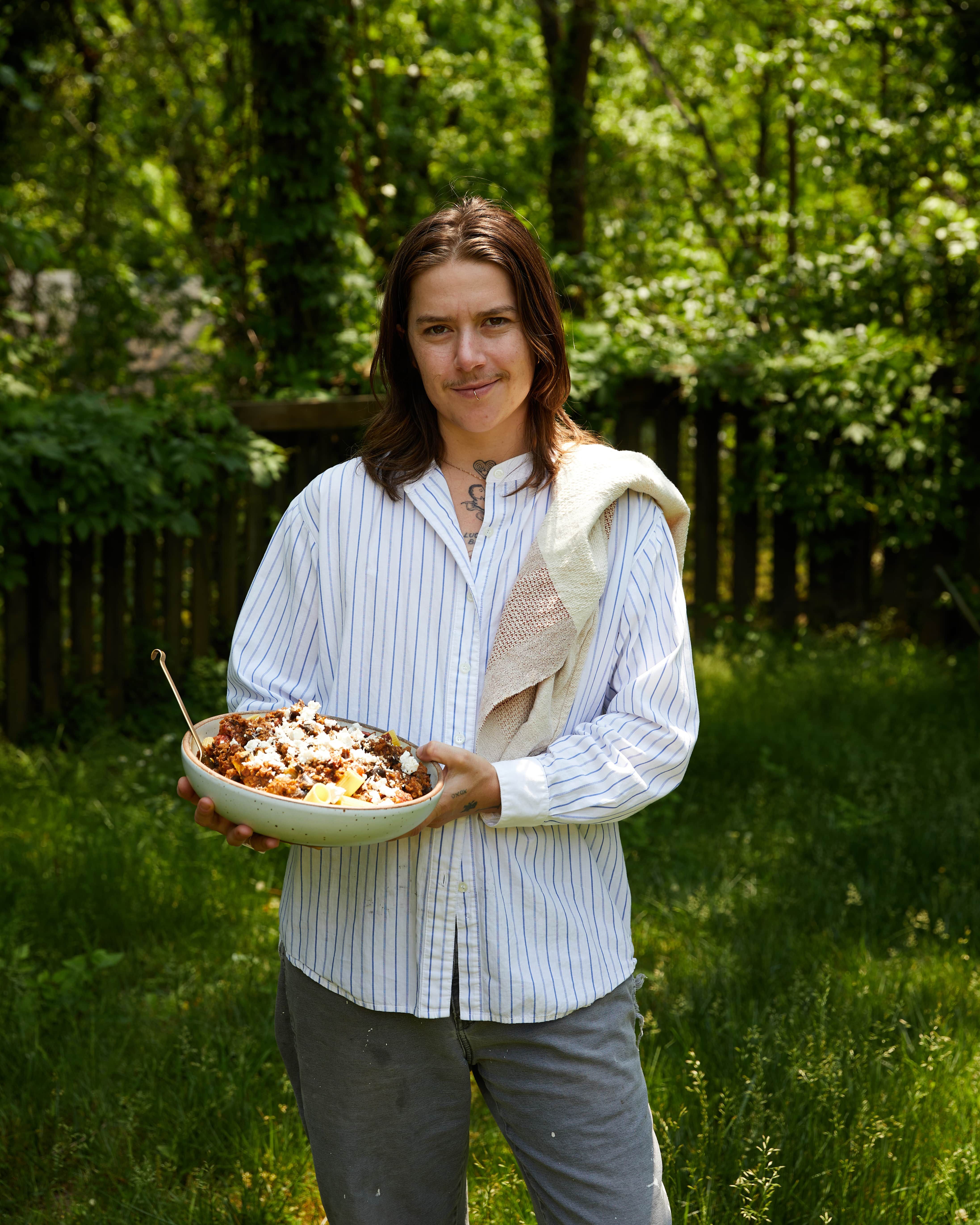 A person stands outside and holds a large serving bowl of rigatoni with a kitchen towel over their shoulder.