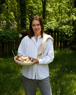 A person stands outside and holds a large serving bowl of rigatoni with a kitchen towel over their shoulder.