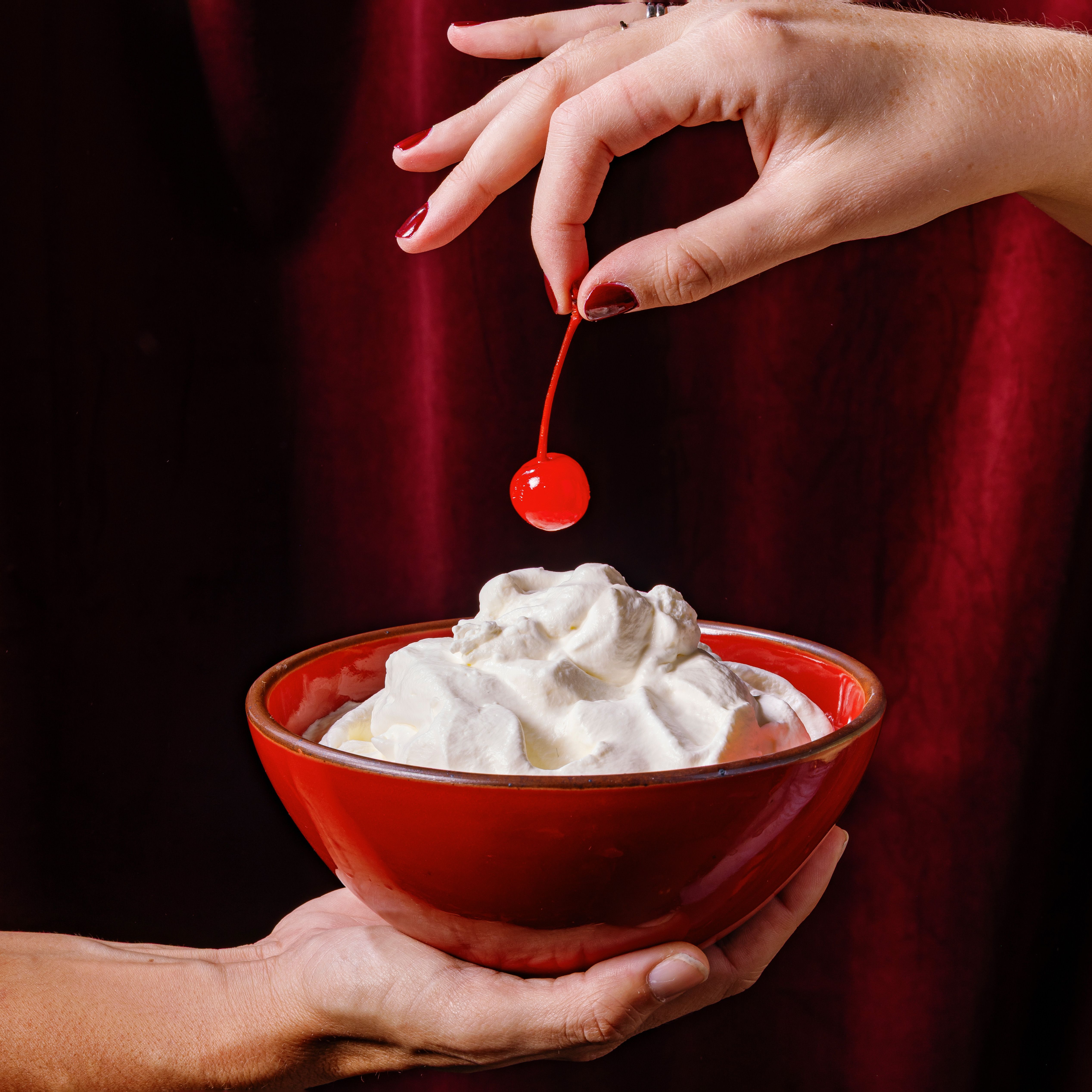 A hand holds out a glossy red ceramic bowl filled with whipped cream, while another hand holds a cherry over it.