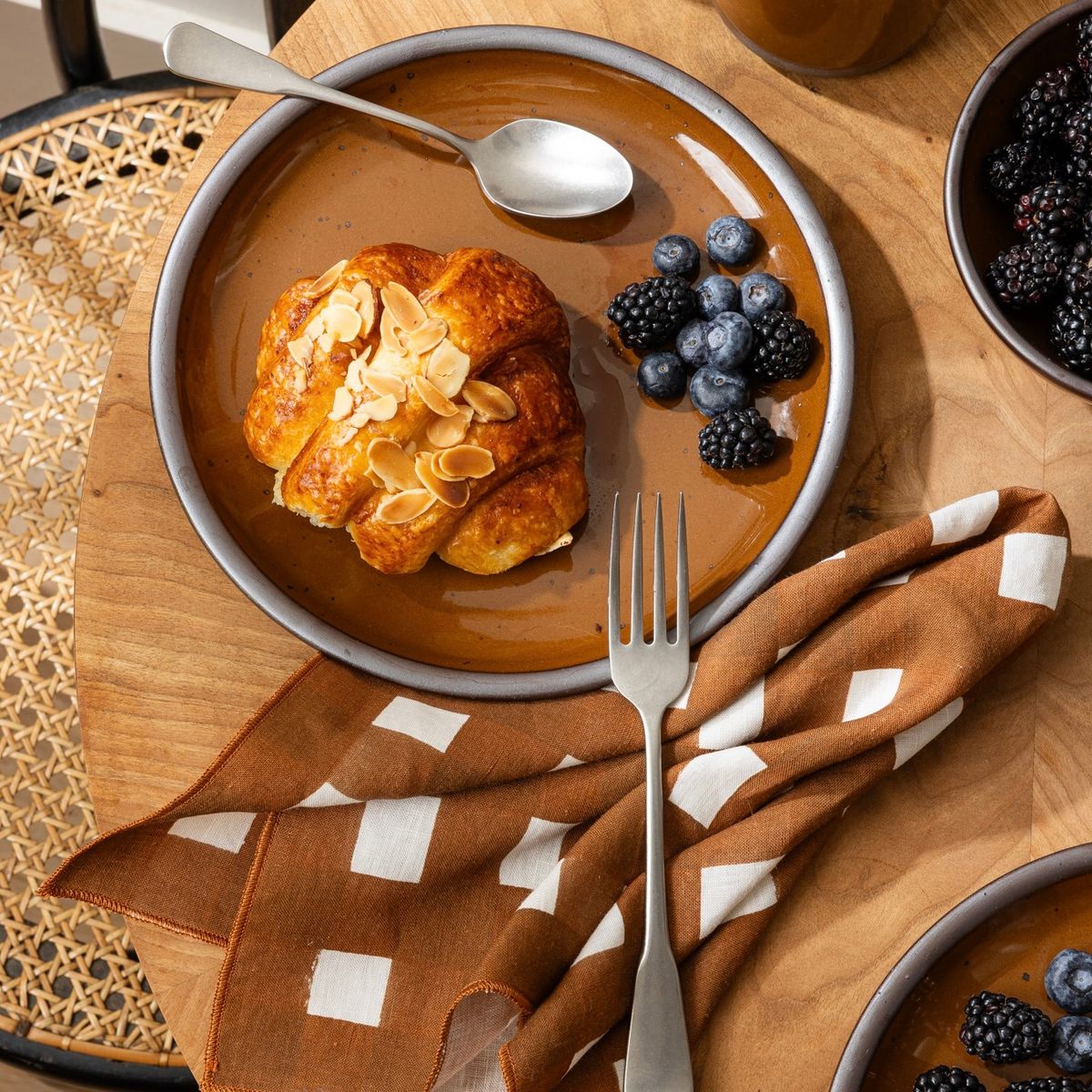 Almond croissant with blueberries and blackberries on a glossy brown ceramic plate with fork, spoon, and patterned napkin.