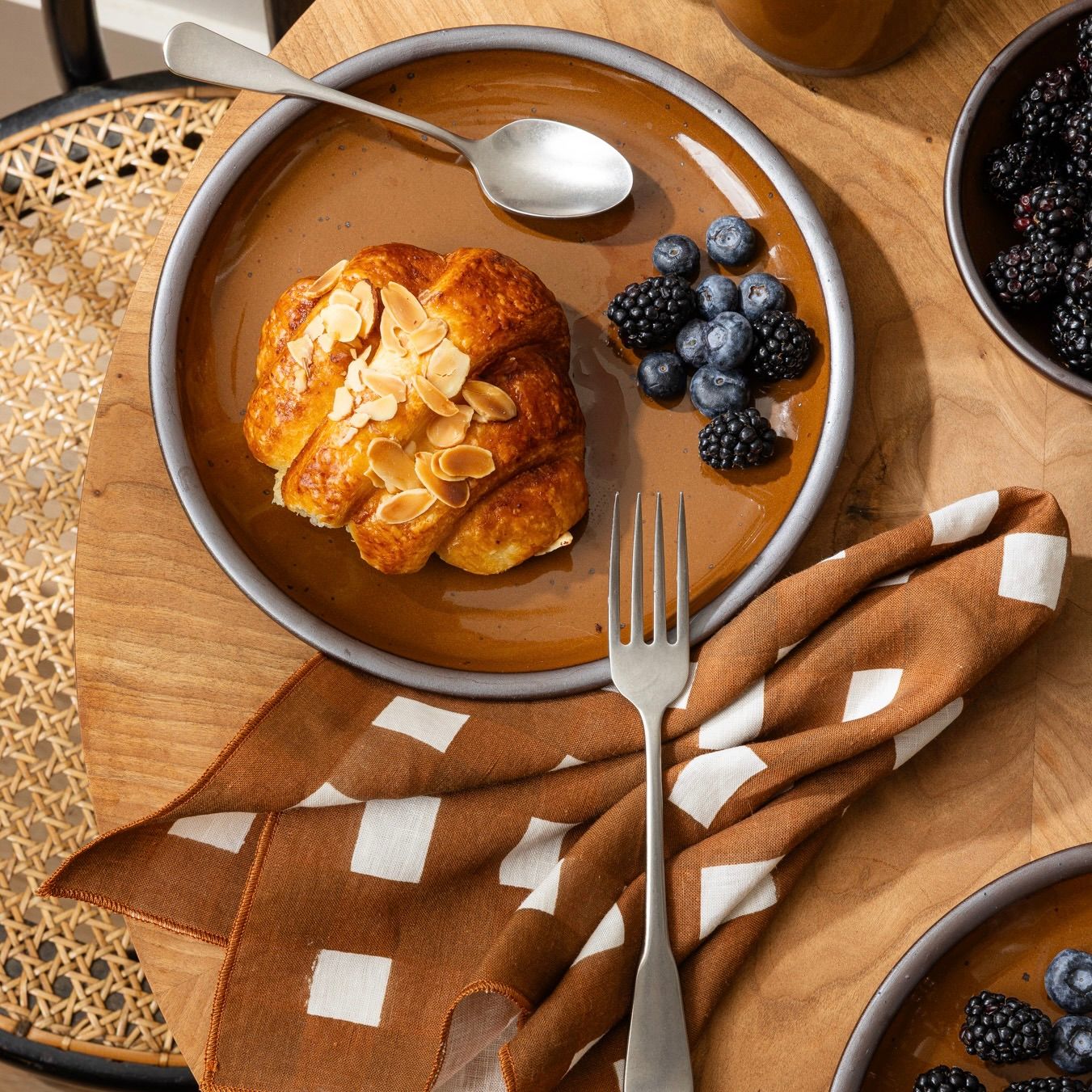 Almond croissant with blueberries and blackberries on a glossy brown ceramic plate with fork, spoon, and patterned napkin.