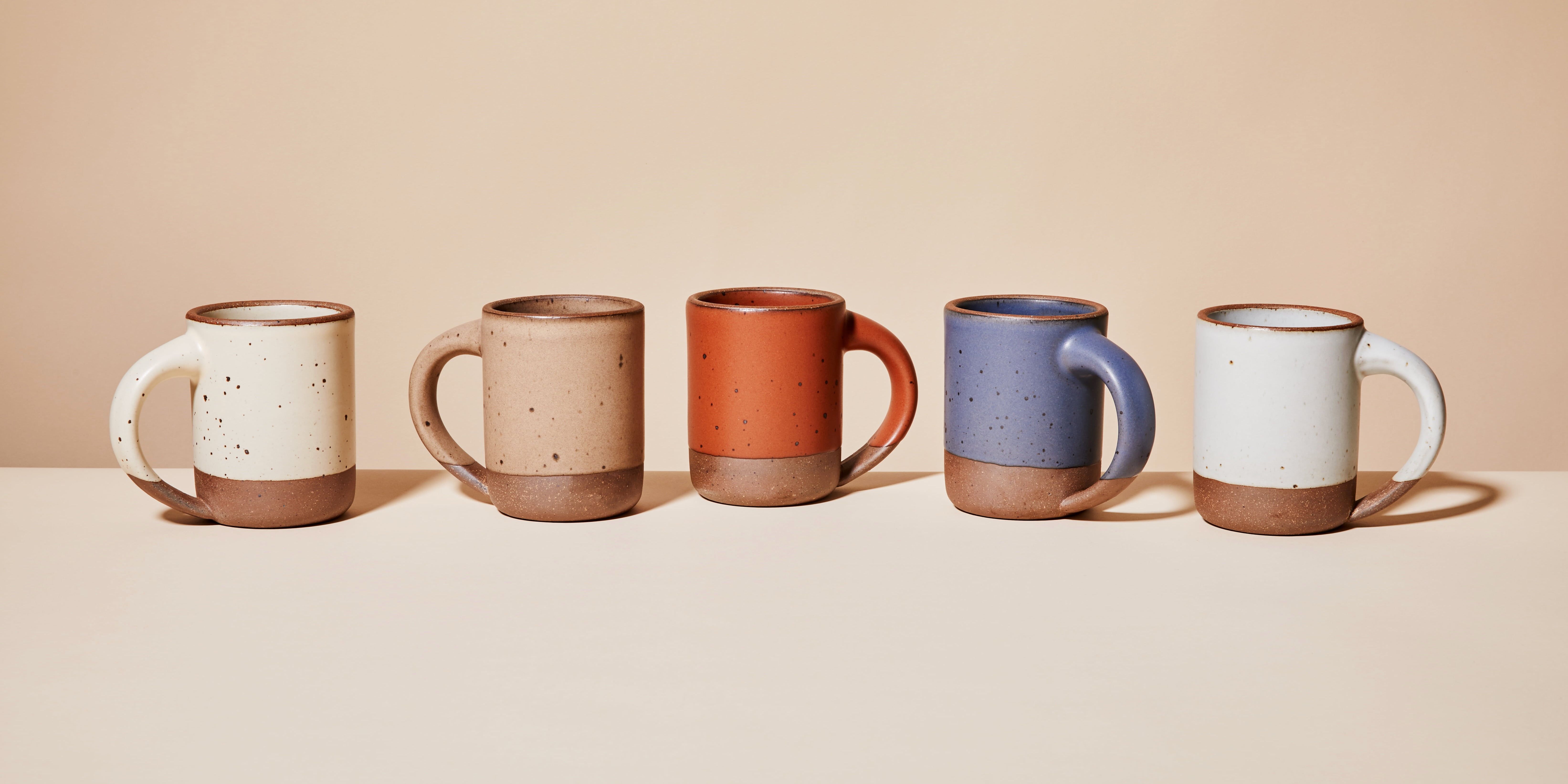 Five ceramic mugs are lined up in a row. Each mug features an unglazed bottom and a ceramic glaze on the body. Each mug has a different color from left to right: cream, muted tan, cool terracotta, muted navy, and cool white.