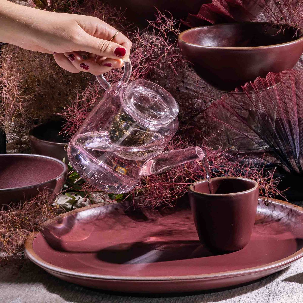 A hand holds a pink glass teapot pouring water into a plum ceramic cup sitting on a plum plate