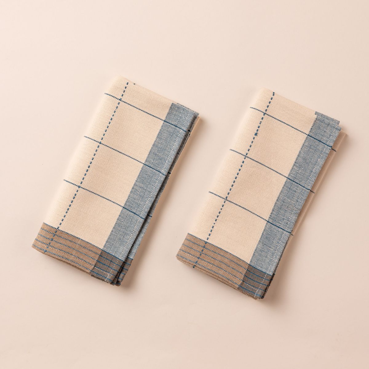  Two natural folded napkins side by side that are designed with turquoise gridlines with light blue and tan striped edging.