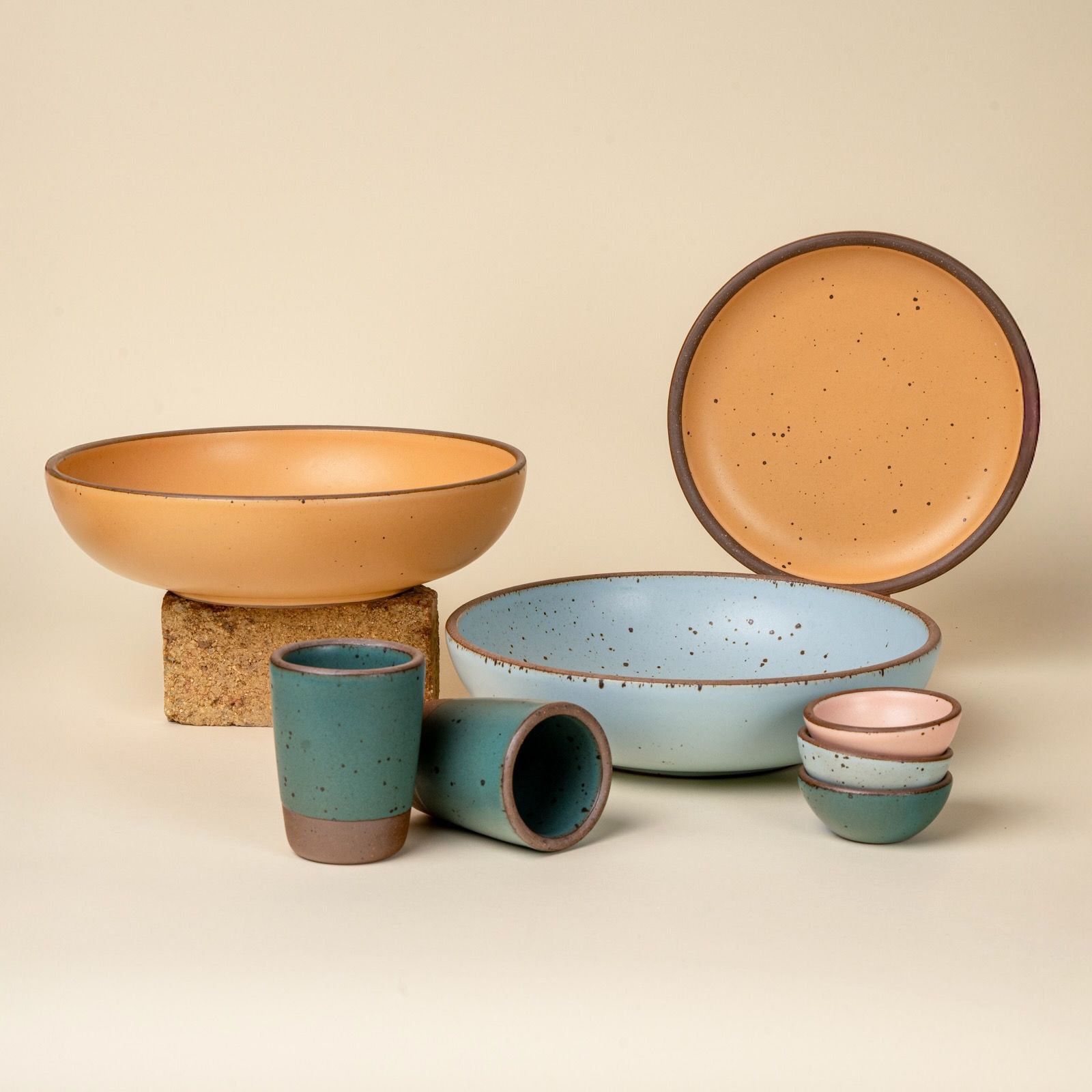 An artful collection of ceramic dinnerware featuring two large serving bowls, one plate, 2 juice cups, and a stack of 3 tiny bowls in teal, baby blue, soft light orange, and baby pink.