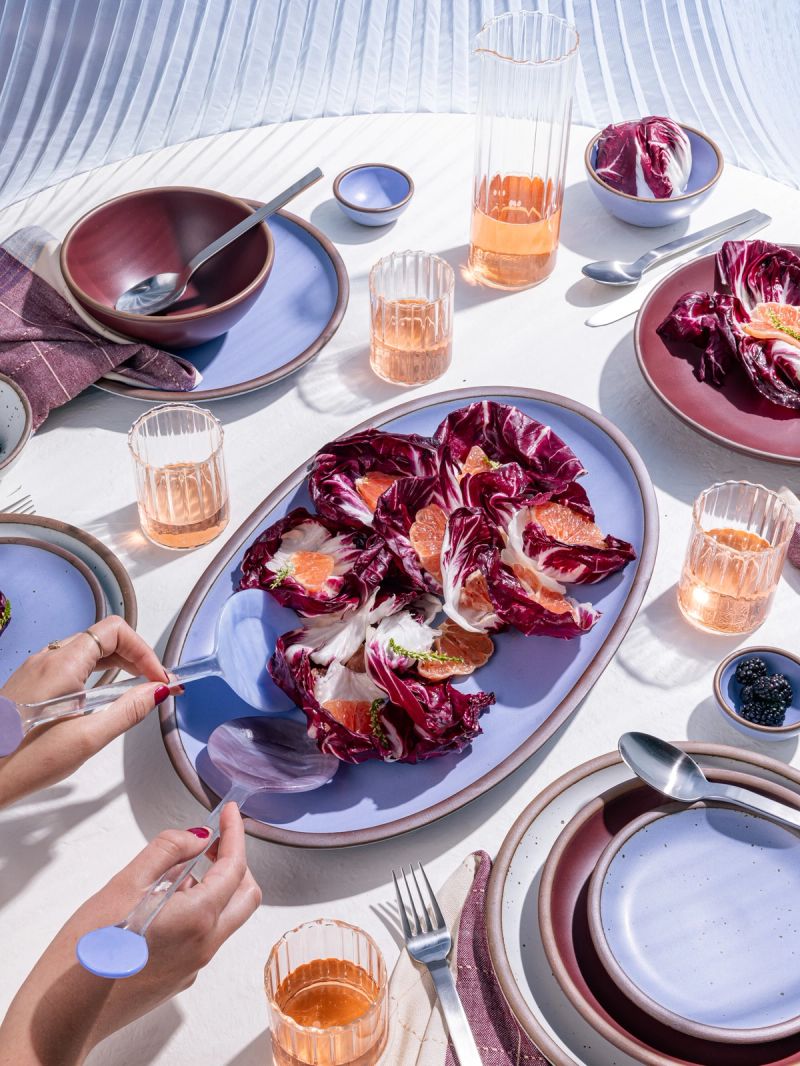 A table filled with ceramic pottery in periwinkle and plum colors, a salad, and glasses filled with orange liquid