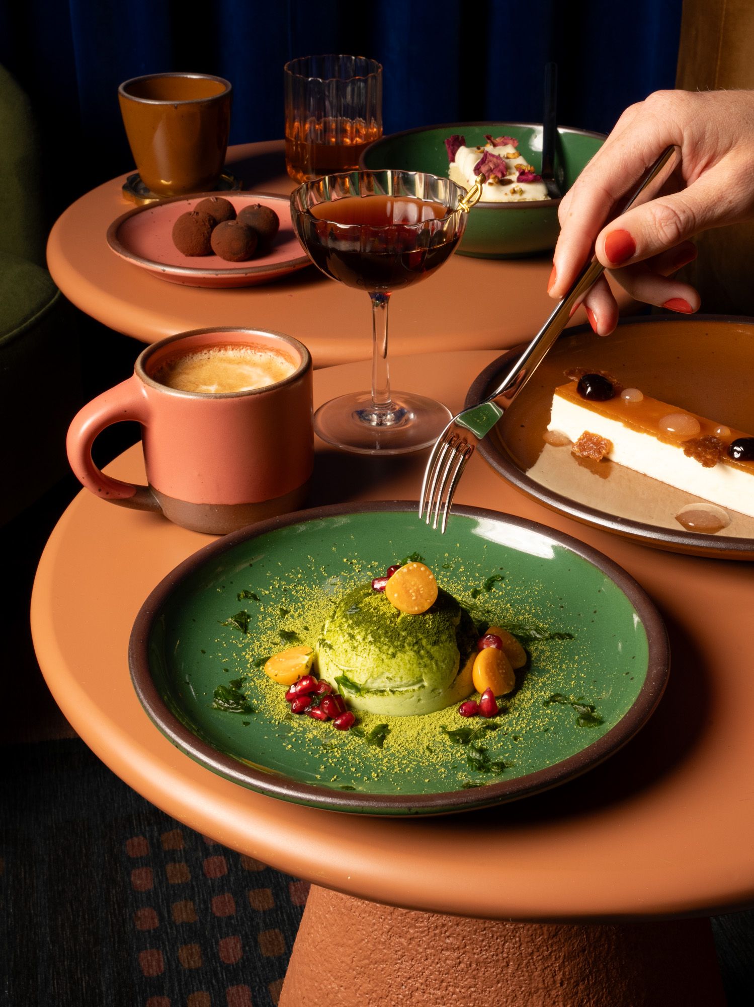 A hand holds a fork above an artful green dessert on a glossy green plate with other ceramic mugs and plates in watermelon pink and glossy brown.