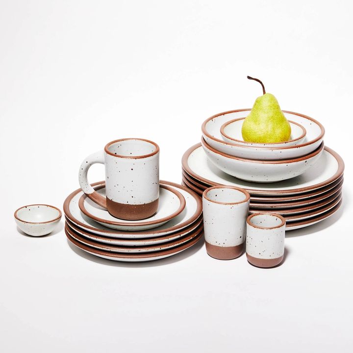 Stack of East Fork pottery: plates, mugs, cups, bowls, cups and bitty bowls in Eggshell, a cool white color, with a pear in one of the top most bowls.