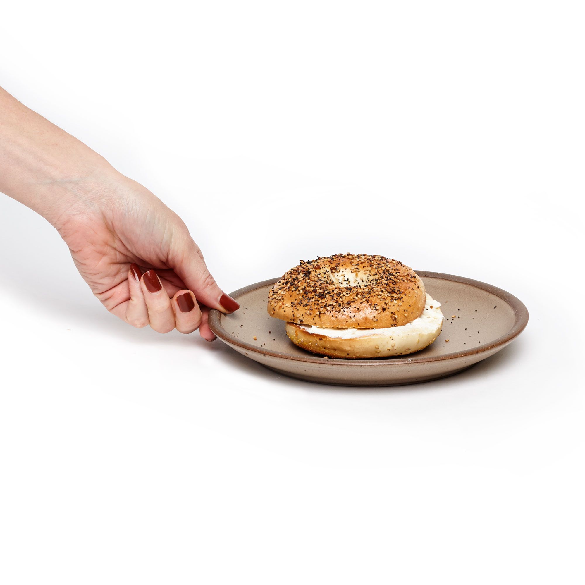 A hand holds a ceramic plate in a warm pale brown color with a sliced bagel on top.