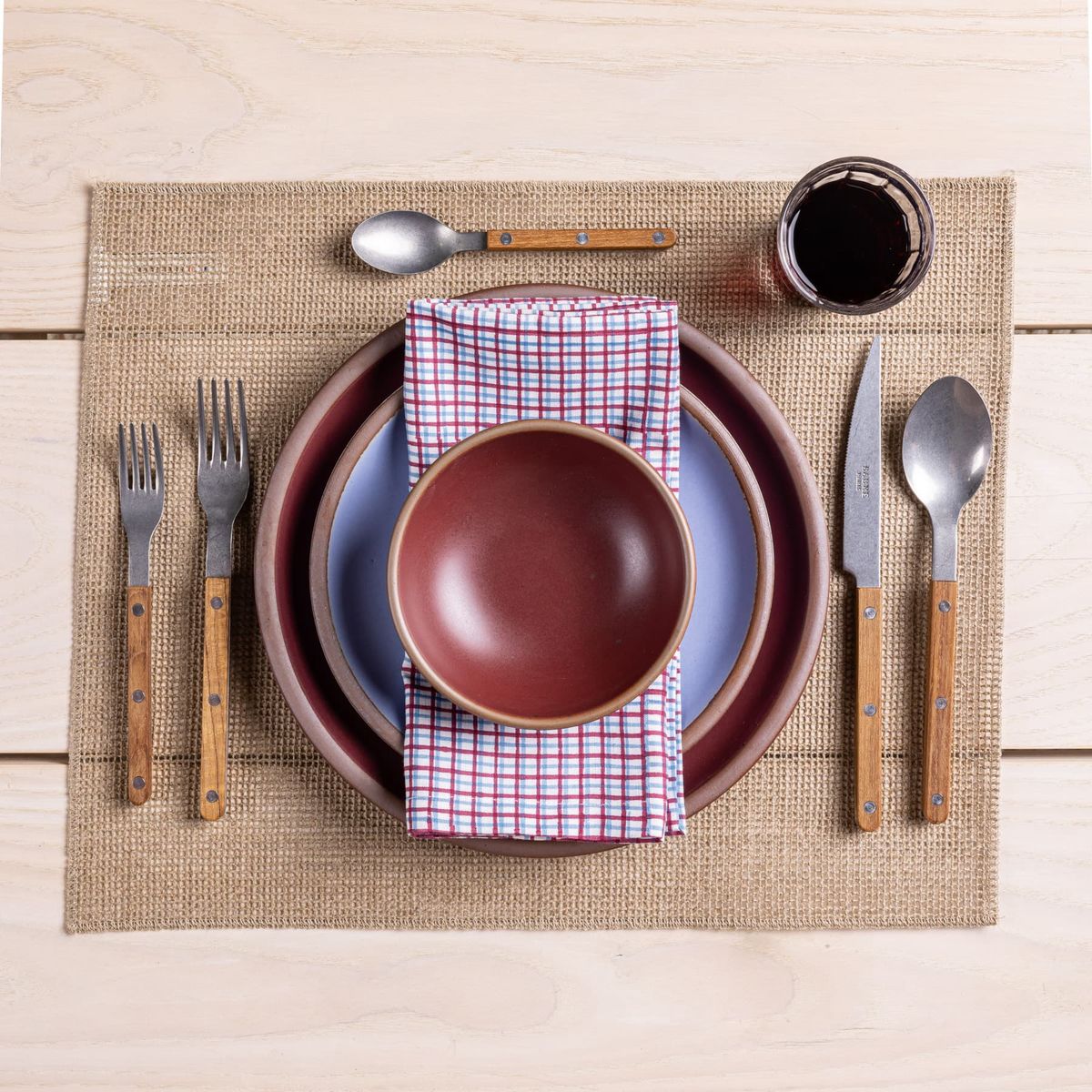 A place setting featuring a grid block printed folded napkin, teak flatware, and plates and bowl in plum and periwinkle colors.